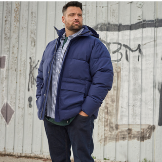 Mand iført mørkeblå North Latitude dynejakke og stribet skjorte står foran slidt grå væg med graffiti.